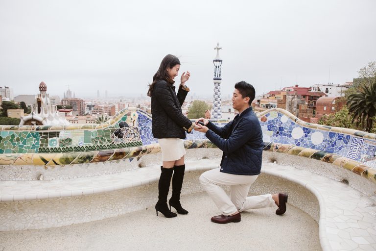 At Park Guell man kneels on one knee proposing to new fiancee.