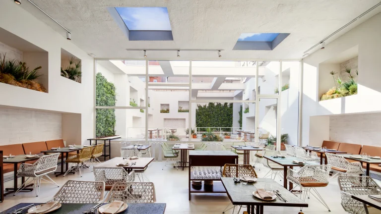 Bright, modern dining room in a fine dining restaurant in Barcelona, with tables facing a window wall that overlooks an open-air courtyard with greenery and sky-view skylights.