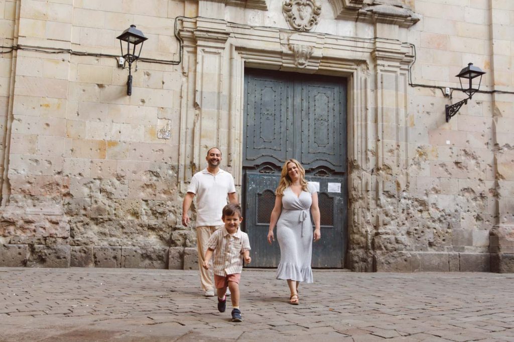 A smiling family of three, with a young boy running ahead of his parents, walks on a cobblestone street in front of an old stone building—one of the 5 iconic locations for a family photoshoot in Barcelona.