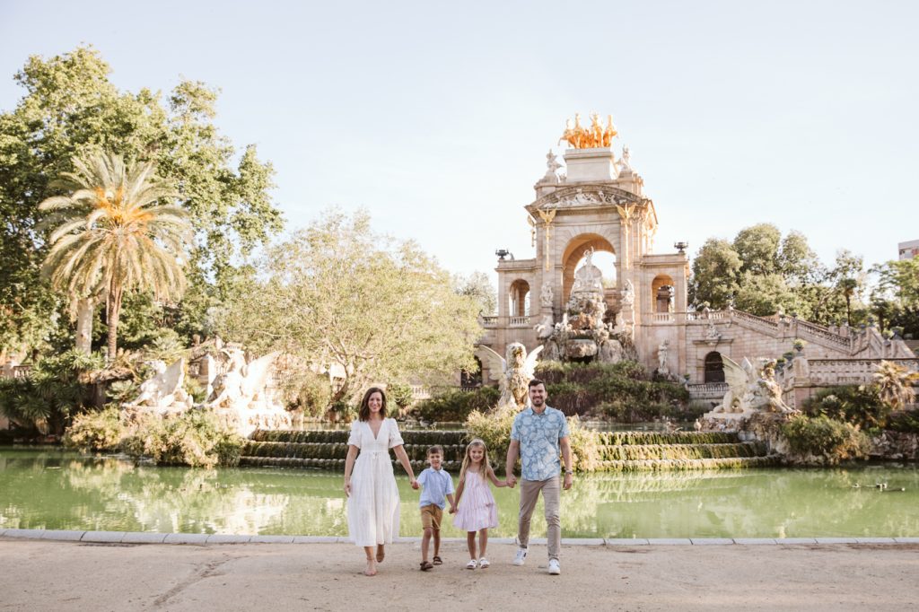 A family of four stands smiling by a pond in front of a grand fountain at Ciutadella Park in Barcelona with statues and lush greenery on a sunny day, capturing the perfect family photoshoot moment.