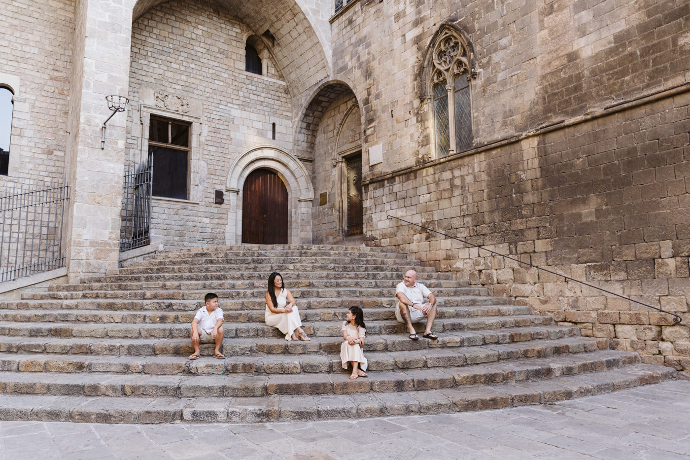 A family photoshoot captures a family of four sitting apart on wide stone steps before an old stone building with arched doors and windows at the Gothic Quarter in Barcelona. Two adults and two children, dressed in light-colored clothing, face each other.