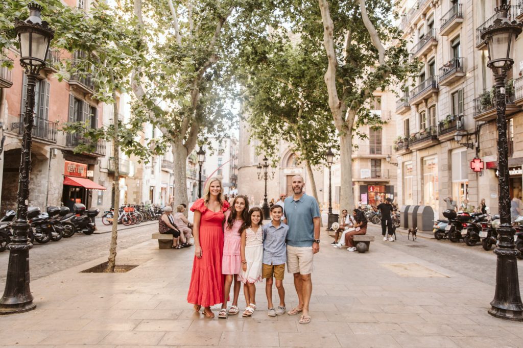 A family of five stands together on a sunny, tree-lined street in a European city for a cheerful family photoshoot, surrounded by historic buildings, motorcycles, and pedestrians. The parents and their three young children smile at the camera.