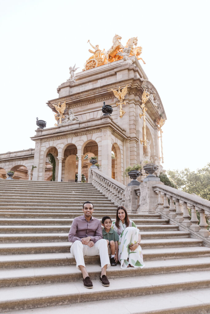 A family of three sits together on wide stone steps in front of an ornate monument with golden statues at the top, under bright daylight—captured perfectly by a skilled family photographer in Barcelona.