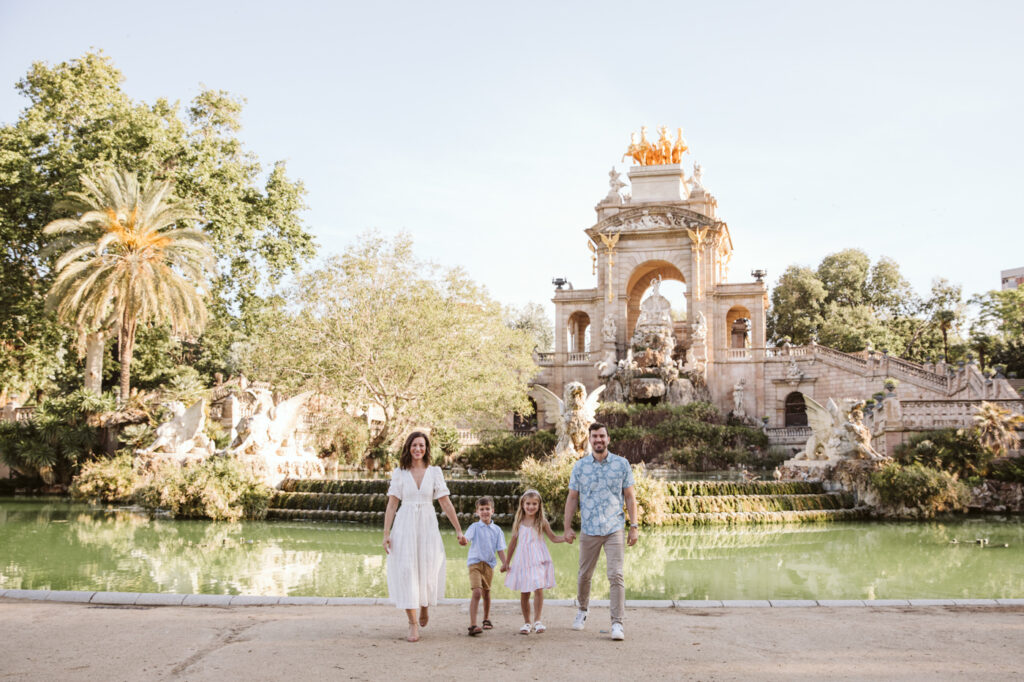 A family of four stands holding hands in front of a large decorative fountain and ornate stone monument, surrounded by lush greenery and water, on a sunny day—captured beautifully by a family photographer Barcelona.