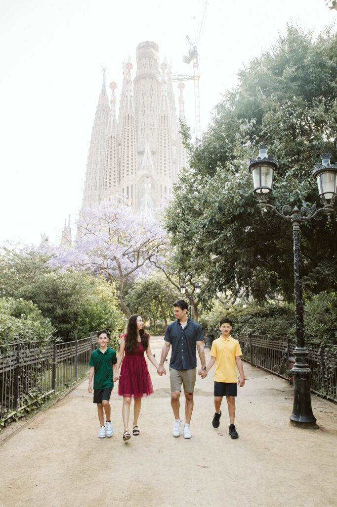 A family of four walks hand in hand down a tree-lined path, the Sagrada Familia basilica visible in the background on a sunny day—perfect for capturing memories with a talented family photographer Barcelona.