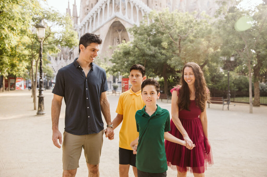 A family of four walks outdoors near Sagrada Familia in Barcelona. The father and two boys hold hands as the mother smiles beside them. Trees and sunlight surround them—a perfect moment for a family photographer in Barcelona to capture.