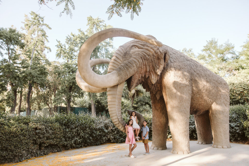 A woman and two children stand in front of a large woolly mammoth statue in a park, surrounded by trees and greenery on a sunny day—an ideal moment for a family photographer Barcelona to capture.