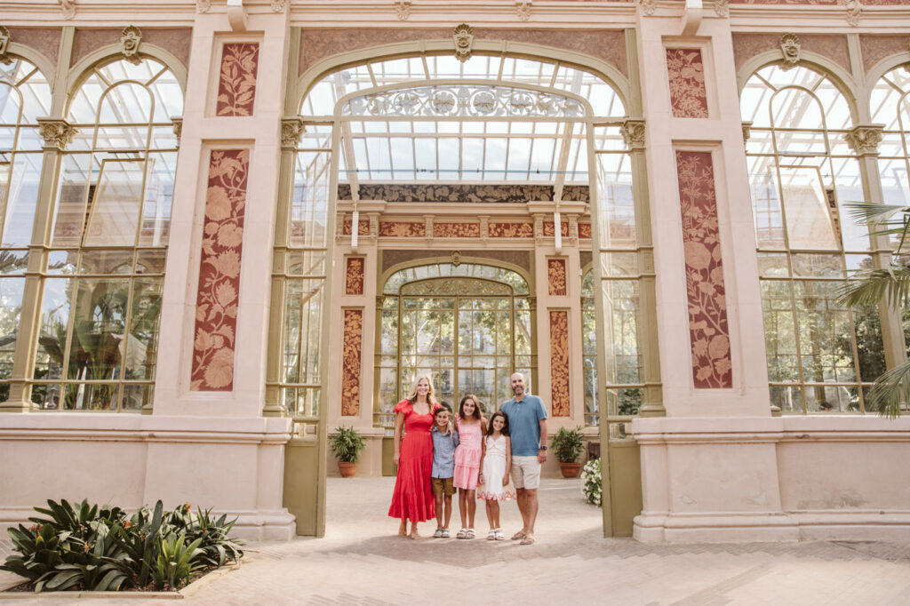 A family of four, two adults and two children, stands smiling in front of a grand glass greenhouse with ornate details and tall windows, captured by a family photographer Barcelona on a sunny day surrounded by lush plants.