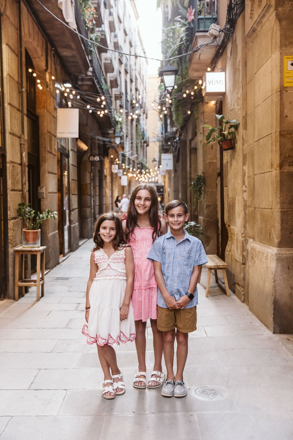 Three smiling children stand together in the middle of a picturesque European street, captured perfectly by a family photographer in Barcelona. Old buildings, potted plants, and string lights create a peaceful, charming atmosphere around them.