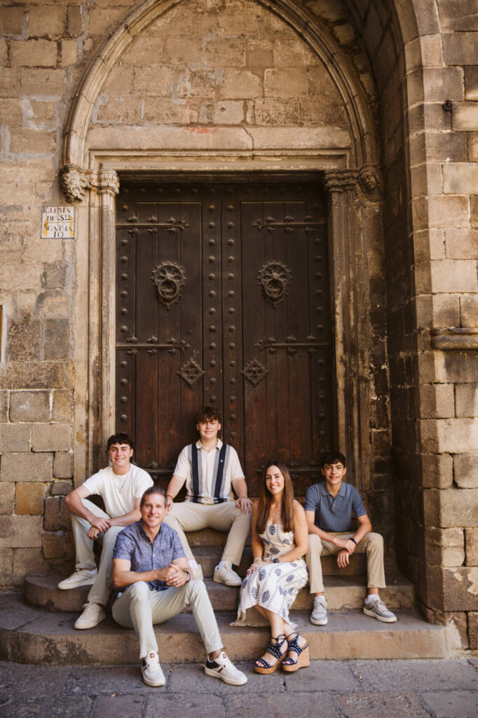 Five young adults pose in front of a large, ornate wooden door with stone archway. Three sit on the steps, while two stand behind them, all smiling and casually dressed. Perfect inspiration for a family photographer in Barcelona seeking a historic setting.