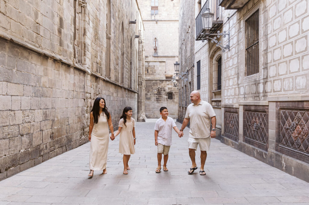 A family of four, dressed in light-colored clothes, walks hand-in-hand down a narrow, historic stone street in Barcelona, smiling and enjoying each other's company—perfect moments for a family photographer Barcelona to capture.