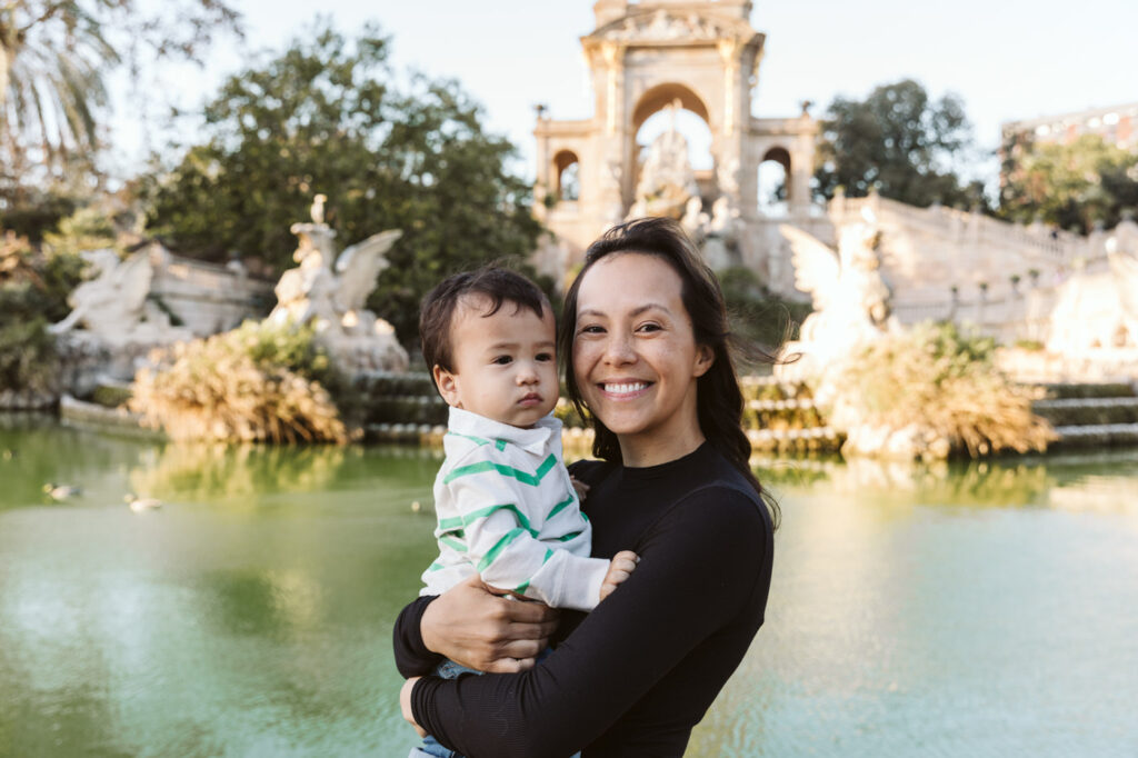 A woman smiles while holding a young child in front of a large ornate fountain and pond in a park on a sunny day, perfectly capturing the joy of the moment—ideal inspiration for any family photographer Barcelona.