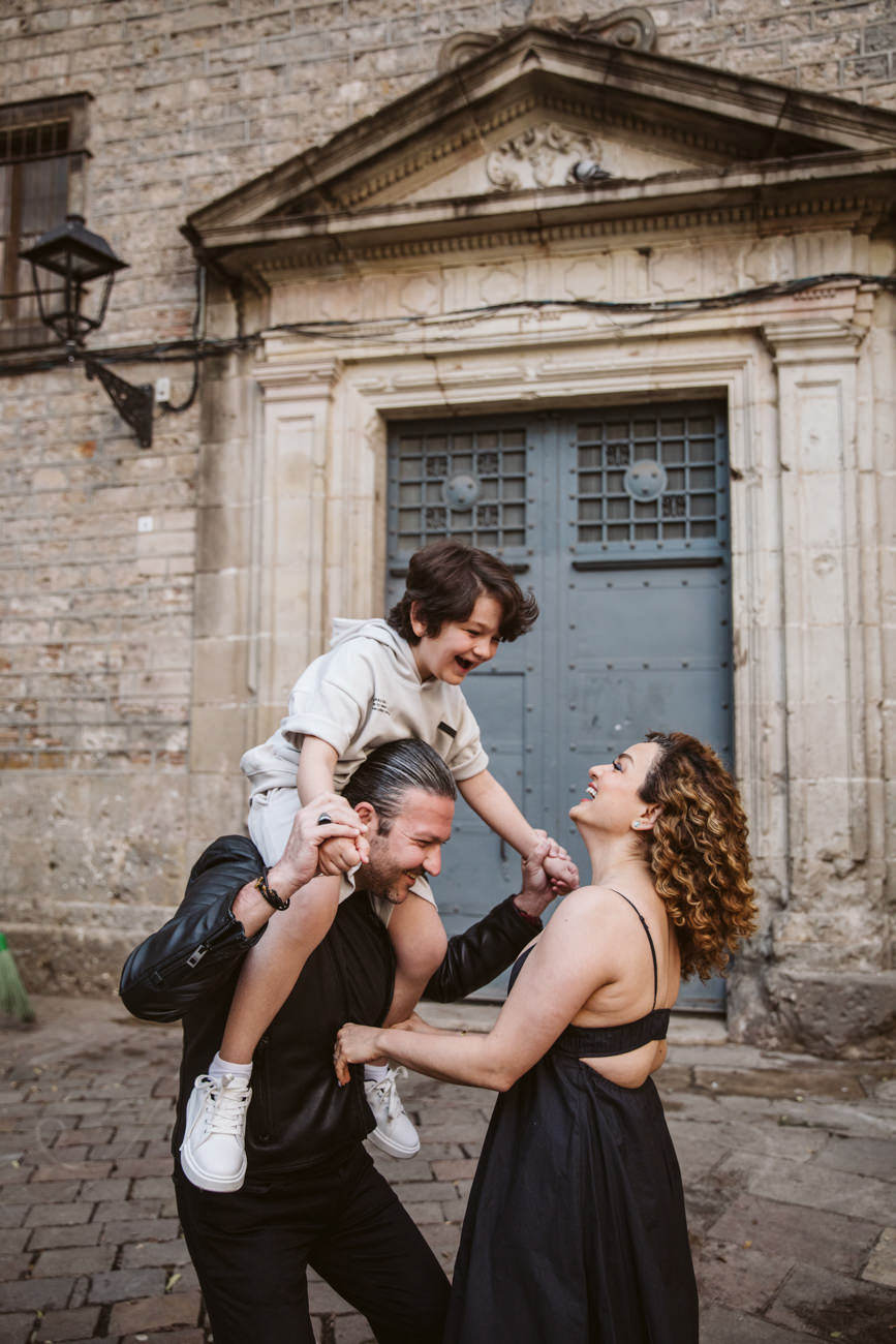 A man carries a smiling boy on his shoulders while a woman in a black dress holds the boy’s hand. Captured by a Barcelona photographer, they all laugh together outside an old stone building with a large blue door.