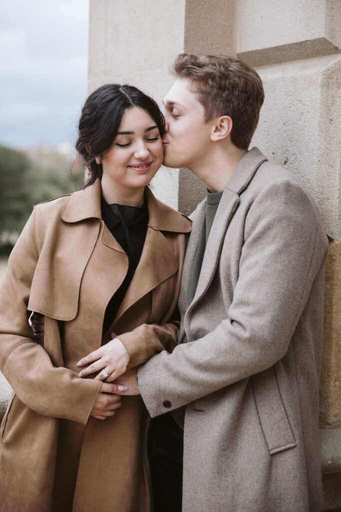 A young man in a light brown coat kisses a smiling woman on the cheek as they stand close together outdoors, both wearing neutral coats against a stone wall—perfect inspiration for barcelona photoshoots pricing ideas.