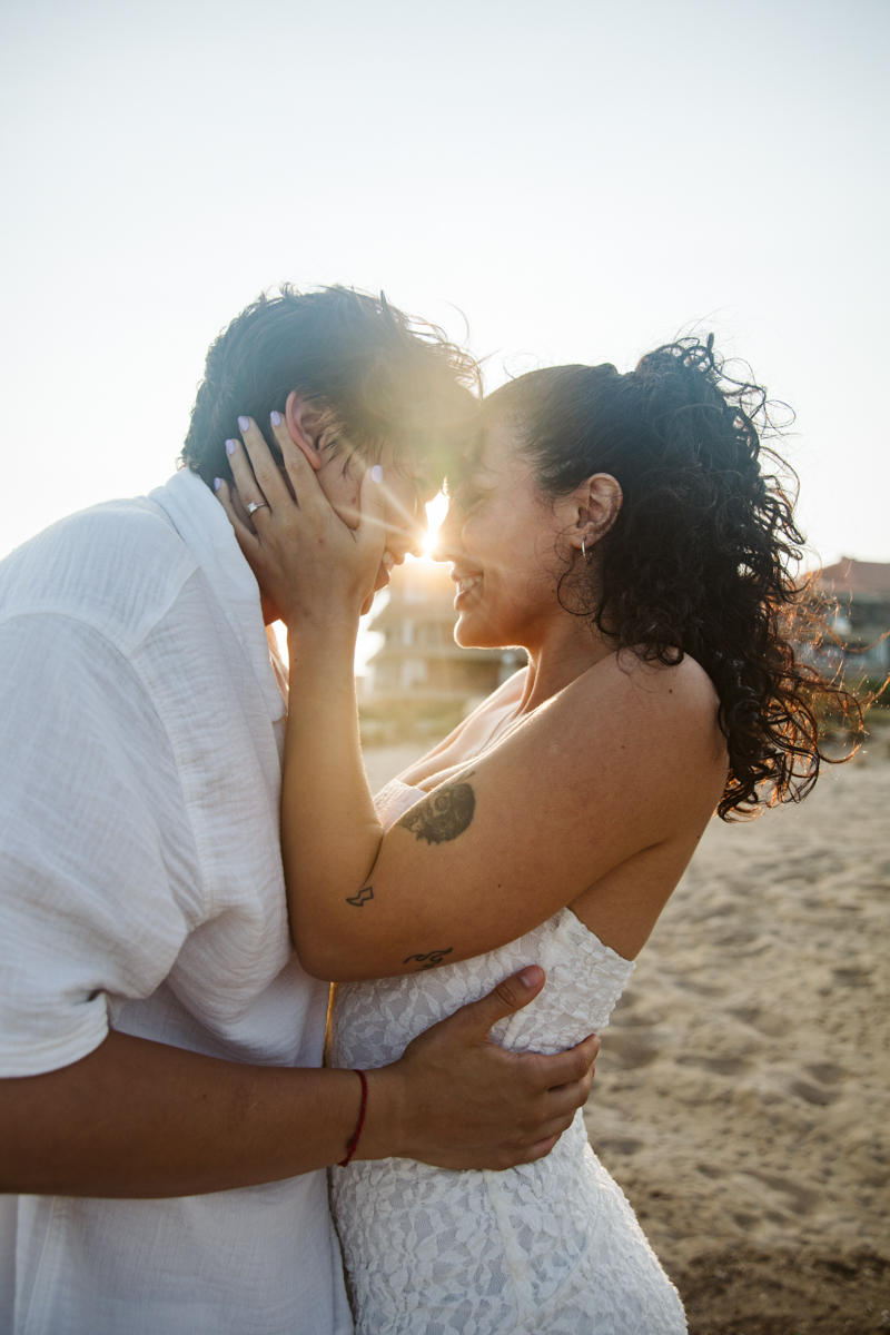 A couple embraces on a sandy beach at sunset, smiling and touching foreheads. Captured by a couple photographer in Barcelona, the woman wears a white dress and holds the man's face as sunlight shines between them.