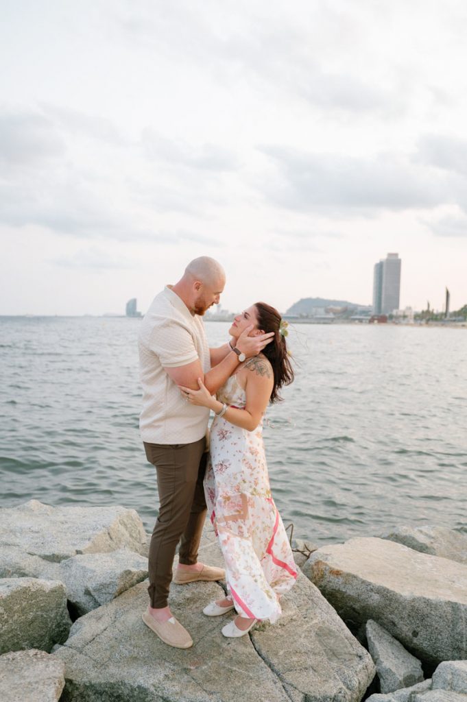 A couple stands on a rocky shore by the sea, gazing into each other’s eyes and smiling. Captured by a couple photographer in Barcelona, the woman wears a floral dress, the man a light shirt, with tall buildings and a cloudy sky in the background.
