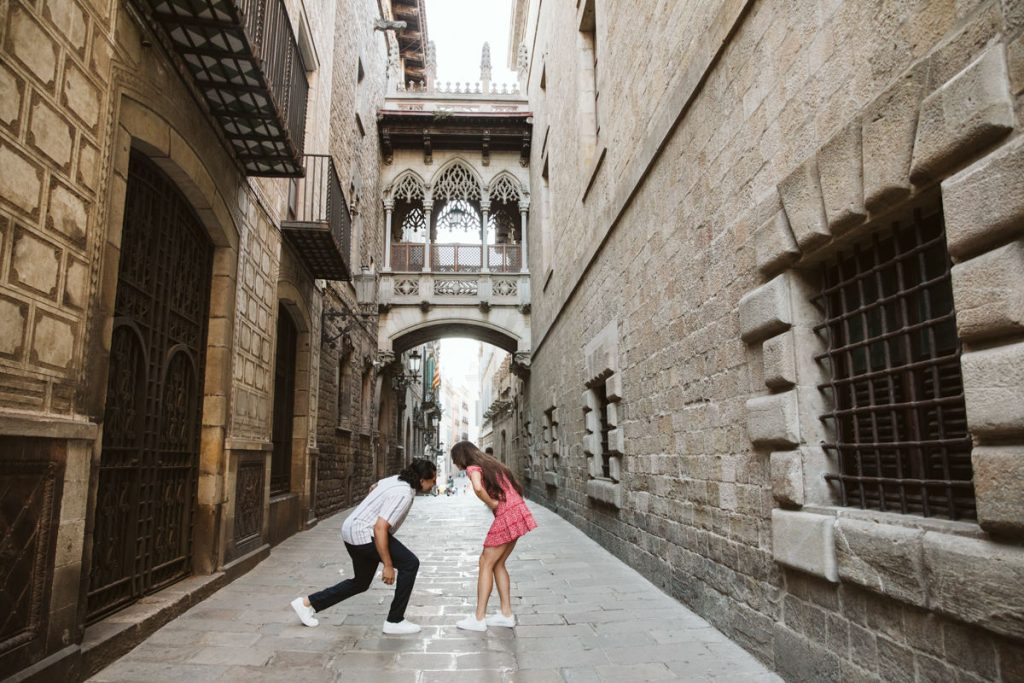 A couple playfully leans forward towards each other in the middle of a narrow, historic stone street lined with buildings featuring arched windows and wrought iron details.