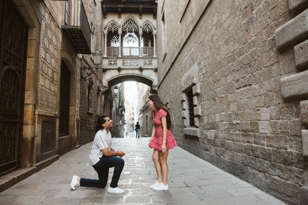 A man kneels on one knee proposing to a smiling woman in a red dress on a cobblestone street, with historic stone buildings and an ornate bridge archway in the background.