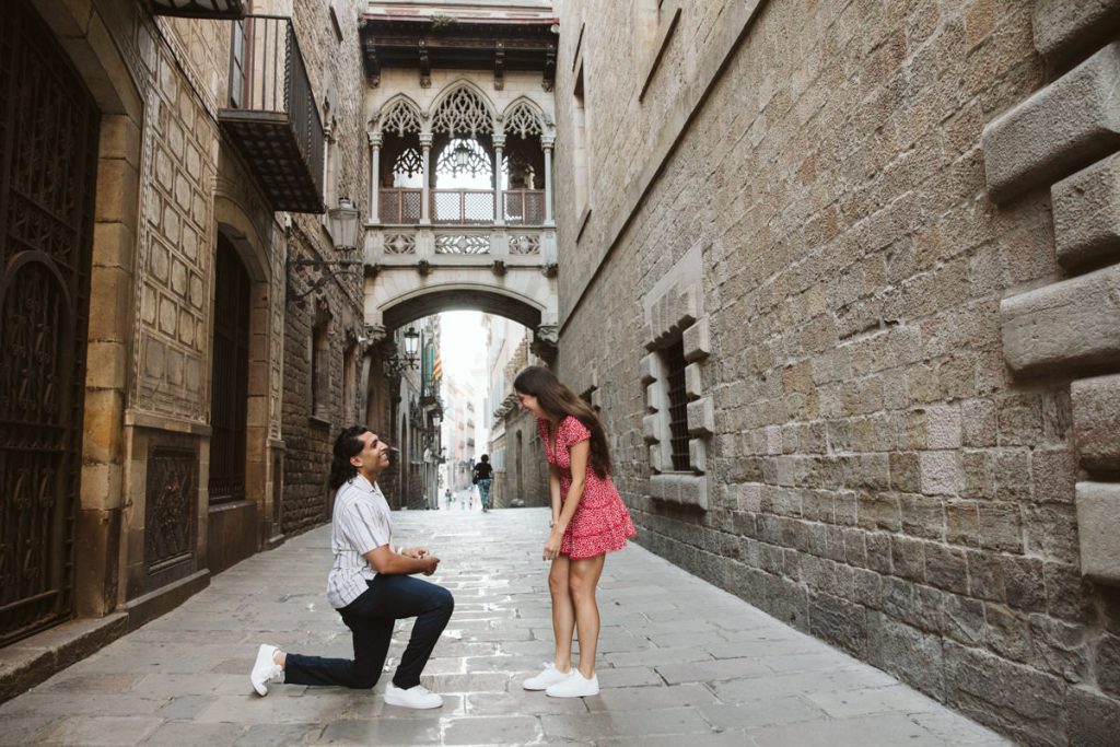 A man kneels on one knee proposing to a surprised woman in a red dress on a narrow, historic stone street with an arched bridge overhead.
