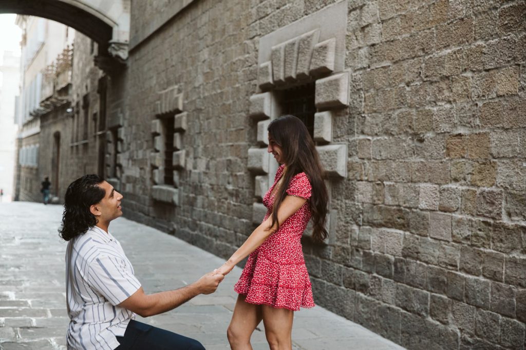 A man kneels on one knee holding a womans hands, proposing to her on a stone-paved street with stone walls; the woman smiles, wearing a red dress, while the man wears a striped shirt.