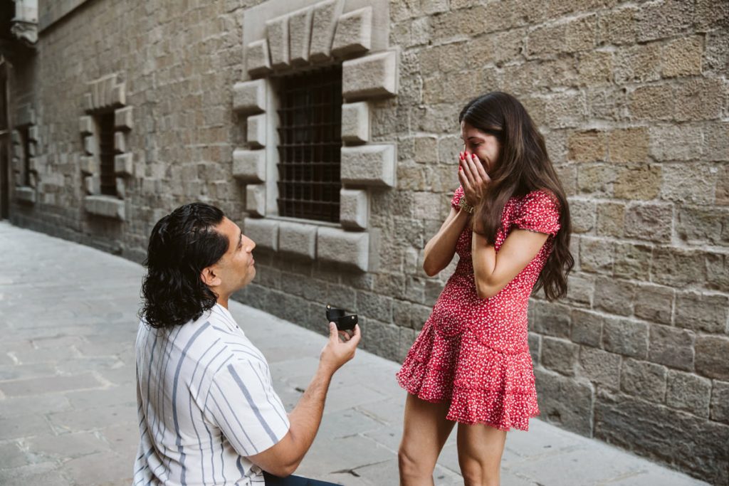 A man kneels on one knee, holding out a ring box to a surprised woman in a red dress. They are on a narrow stone street with historic buildings in the background.