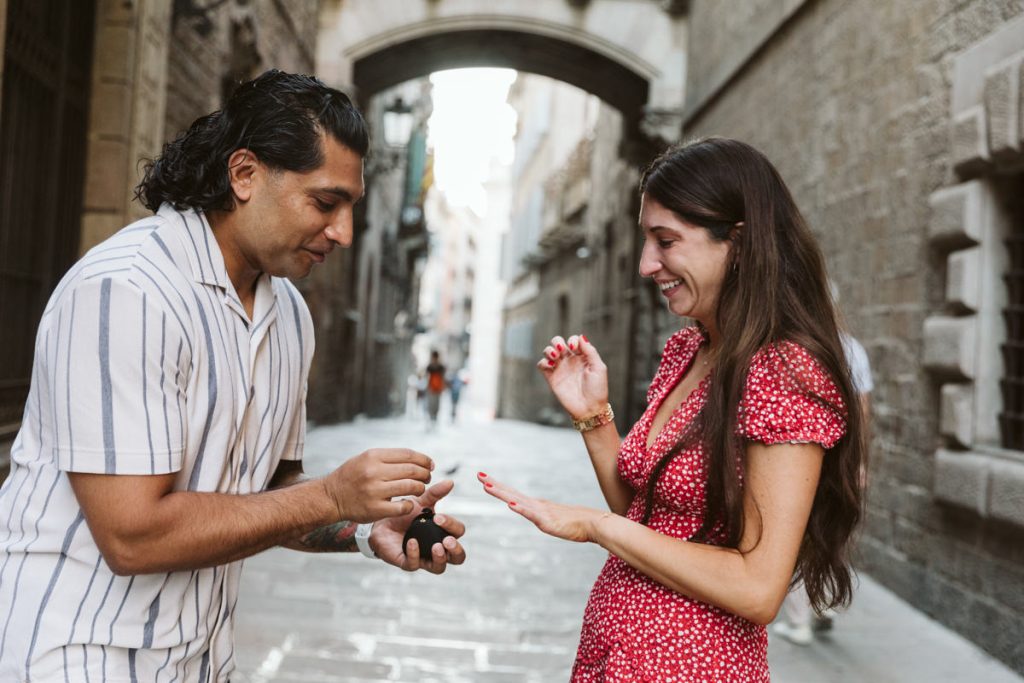 A man in a striped shirt proposes with a ring to a smiling woman in a red dress on a charming, narrow street with stone buildings. The woman looks excited and happy, holding out her hand.