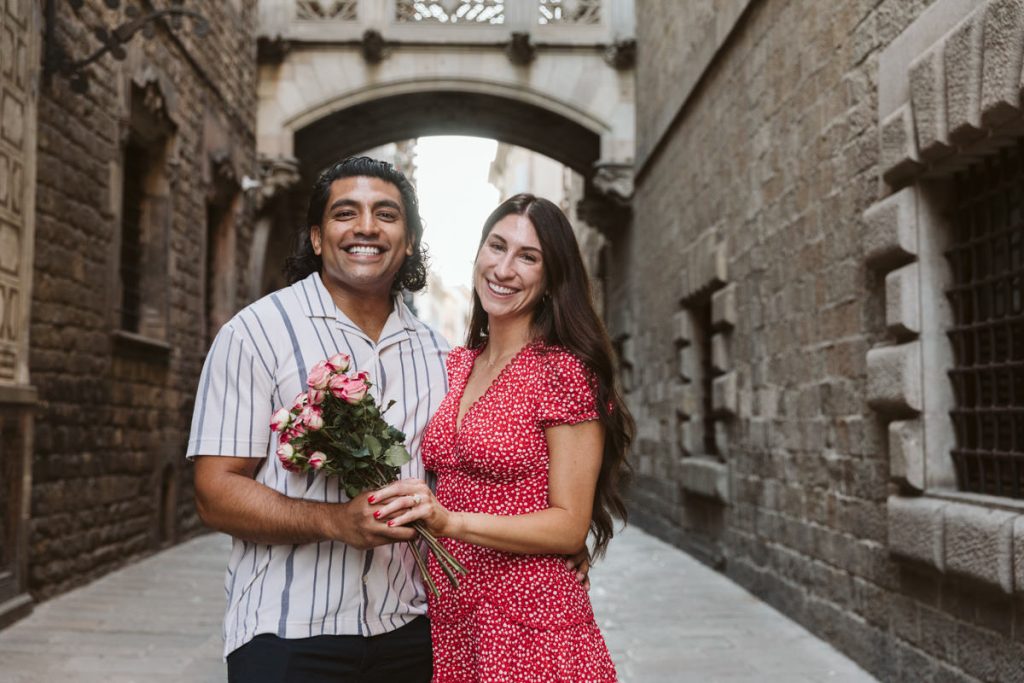 A smiling couple stands close together in a narrow stone street, holding a bouquet of pink roses. The woman wears a red dress with white dots; the man wears a striped shirt. An arched bridge spans the alley behind them.