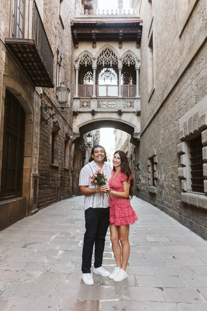 A smiling couple stands close together in a narrow, historic stone street, holding flowers. Ornate architecture and a decorative bridge appear in the background. The woman wears a red dress; the man wears a striped shirt and dark pants.