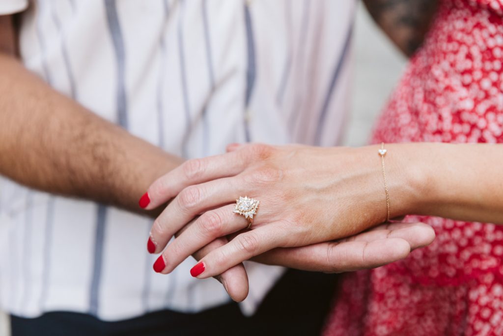 A close-up of two hands, one resting over the other. The top hand has red nail polish, a delicate bracelet, and wears a sparkly engagement ring with a large center stone. The person wears a red patterned dress.