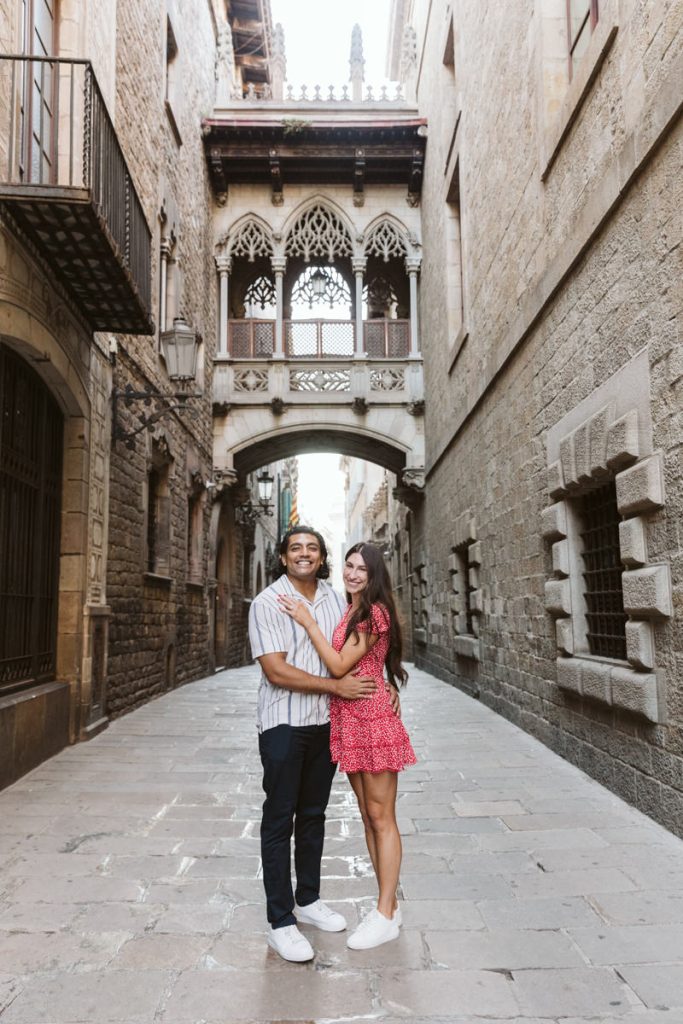 A smiling couple stands close together on a cobblestone street, surrounded by old stone buildings with balconies and a decorative archway above them. The woman wears a red dress, and the man wears a striped shirt and dark pants.