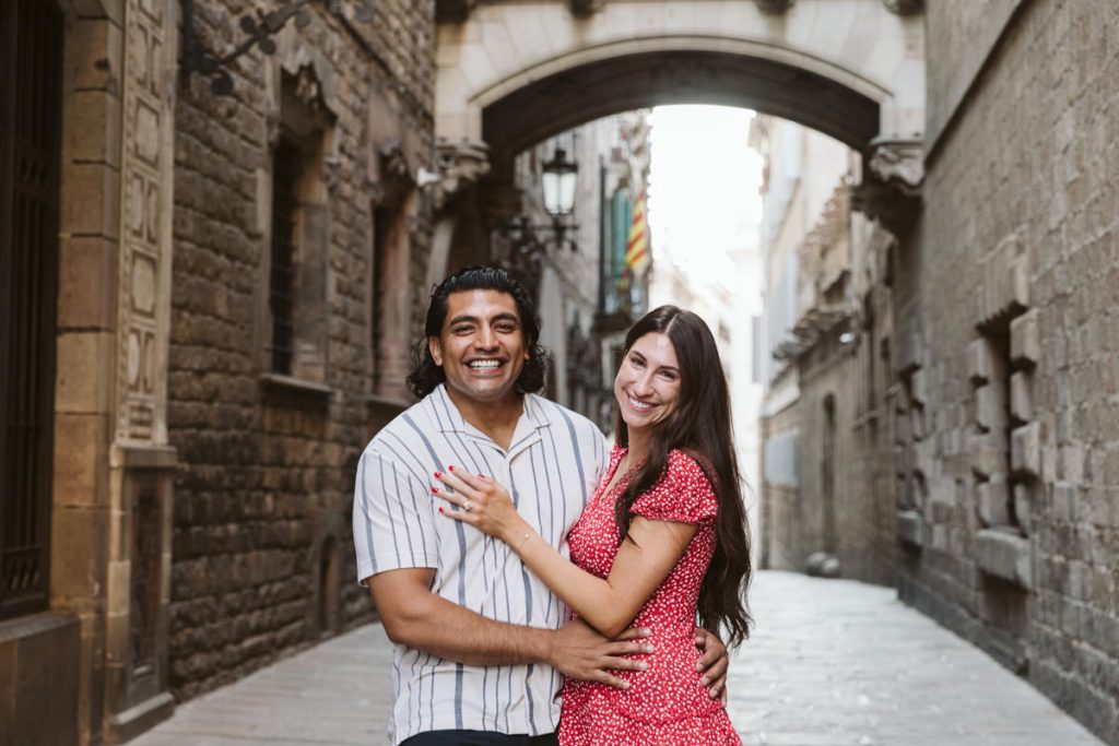 A smiling couple stands close together on a narrow cobblestone street, surrounded by old stone buildings and an archway. The man wears a striped shirt; the woman wears a red dress with white spots.