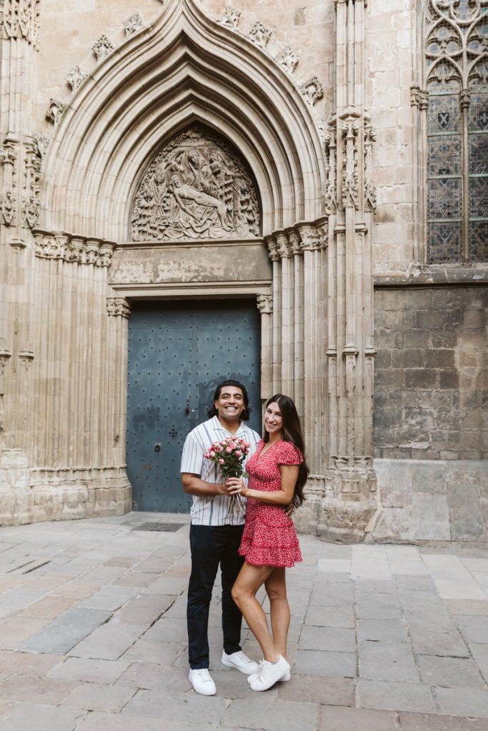 A smiling couple stands close together in front of an ornate, historical stone building with a large arched doorway. The man holds a bouquet of flowers, and the woman wears a red dress and white sneakers.