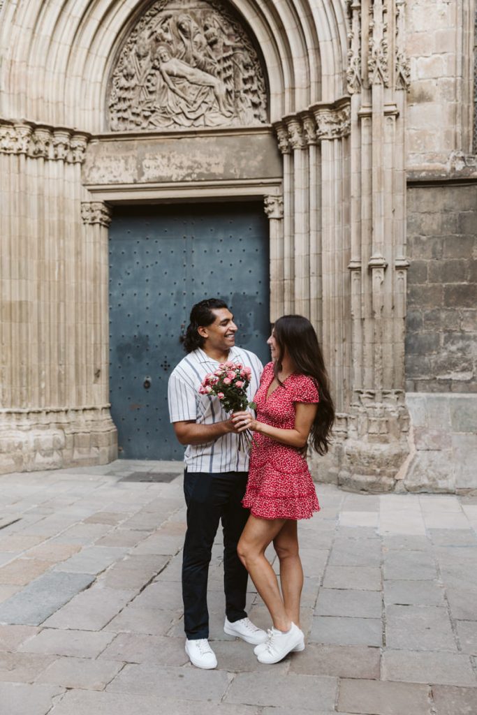A man and woman smile at each other, holding a bouquet of flowers, standing in front of an ornate, arched wooden door set in a stone building. The woman wears a red dress; the man wears a striped shirt and dark pants.