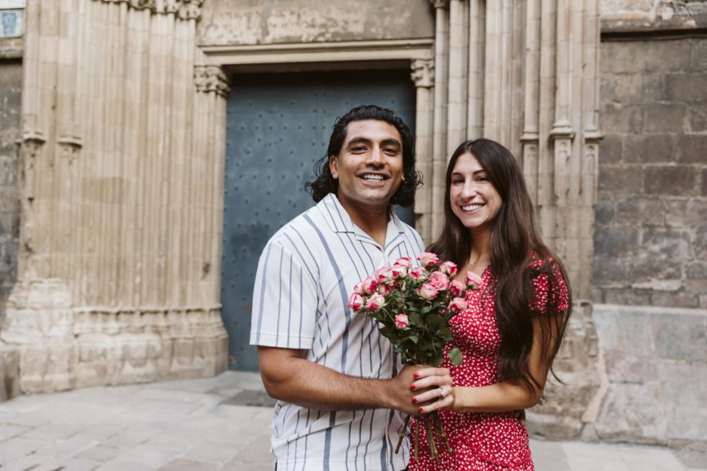 A smiling couple stands outside a historic building. The man holds a bouquet of pink roses, and the woman, wearing a red dress with white dots, stands beside him, both looking at the camera.