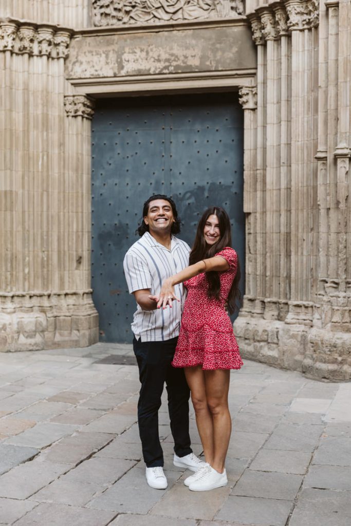 A smiling couple stands in front of an ornate stone doorway. The woman, wearing a red dress, holds out her hand to display a ring, while the man stands beside her with his arm around her waist.