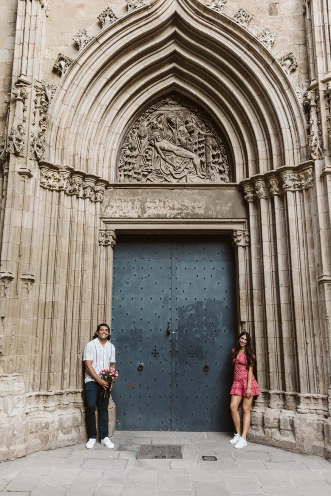 A man and a woman stand apart in front of a large, ornately carved Gothic archway and blue doors on a stone building. The man holds flowers and wears a striped shirt; the woman wears a red dress.