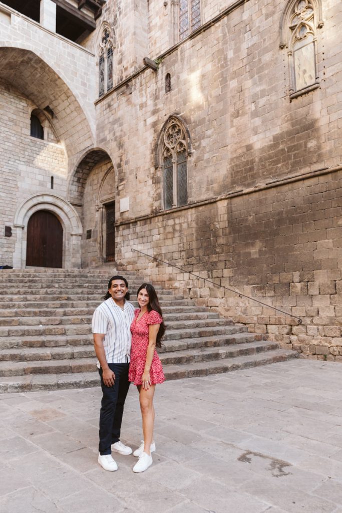 A smiling couple stands together on a stone courtyard in front of historic stone steps and an old building with arched doors and tall windows. The woman wears a red dress; the man wears a striped shirt and dark pants.