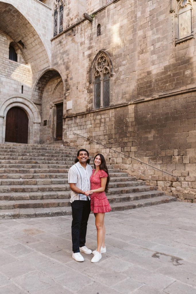 A couple stands holding hands and smiling on a stone courtyard in front of historic, arched architecture with wide steps and tall windows. The man wears dark pants and a striped shirt; the woman wears a short red dress and white shoes.