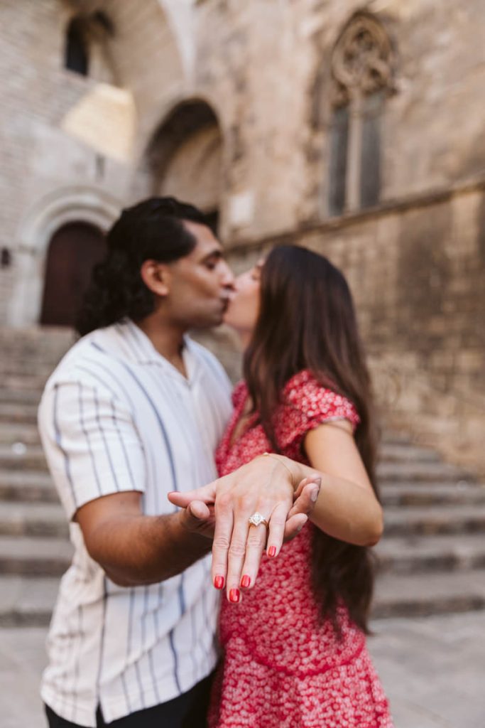 A couple kisses as the woman extends her hand toward the camera, displaying an engagement ring. They stand on stone steps with an old building in the background.