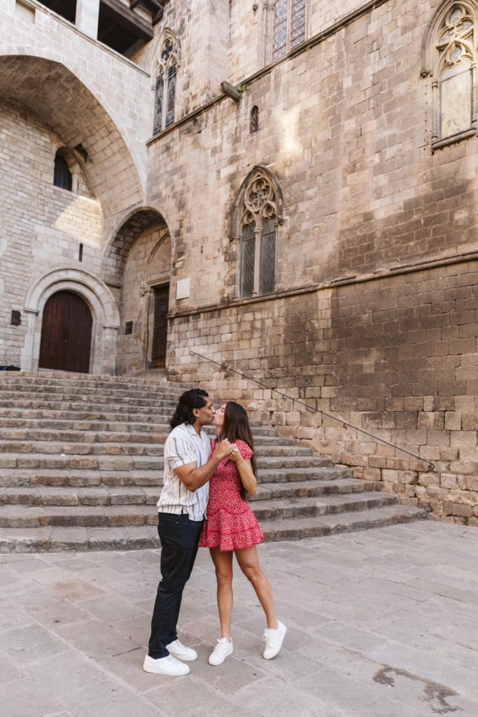 A couple stands on stone steps outside a historic building, sharing a kiss. The woman wears a red dress and white sneakers; the man wears a striped shirt, dark pants, and white shoes. The building has arched doorways and windows.
