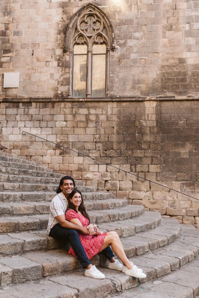 A couple sits together on stone steps in front of an old brick wall with a large arched window. The man hugs the woman from behind as they both smile at the camera.