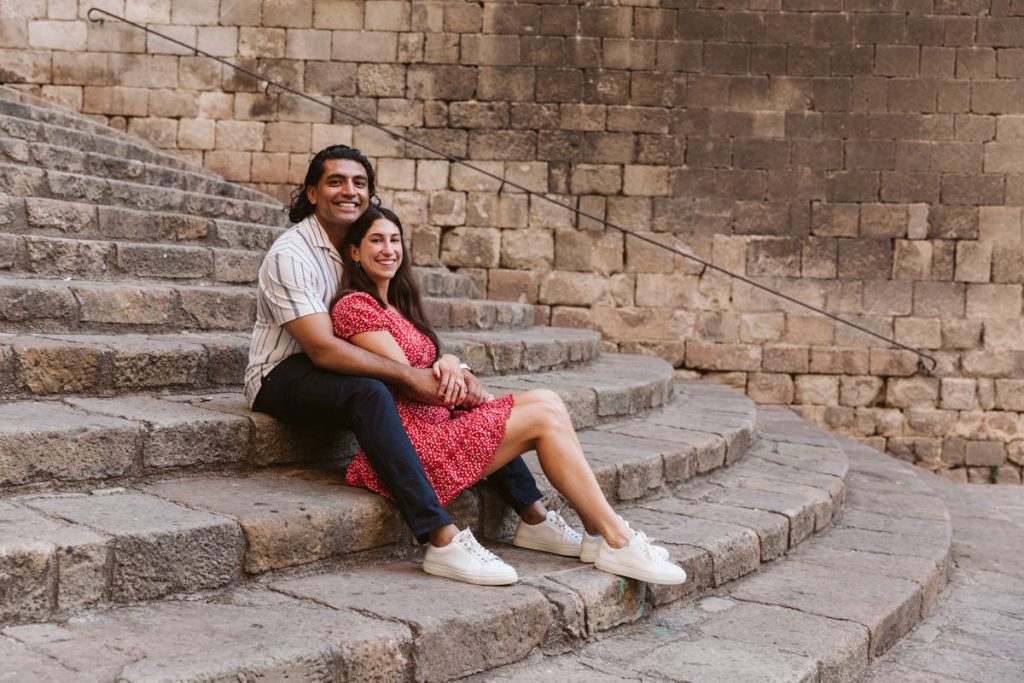 A couple sits together on stone steps, smiling at the camera. The woman wears a red dress and white sneakers, while the man is dressed in a striped shirt and dark pants. They are surrounded by old stone walls.