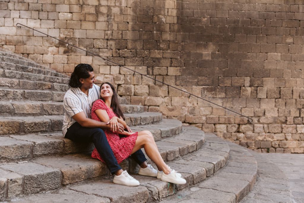 A couple sits on stone steps, smiling and embracing. The woman wears a red dress and white sneakers, while the man wears a striped shirt and dark pants. Behind them is a large stone wall.