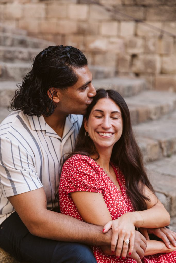 A man with long dark hair kisses a smiling woman on the forehead as they sit on stone steps. The woman, wearing a red dress with white dots, has her eyes closed and rests her hand on his arm, showing an engagement ring.