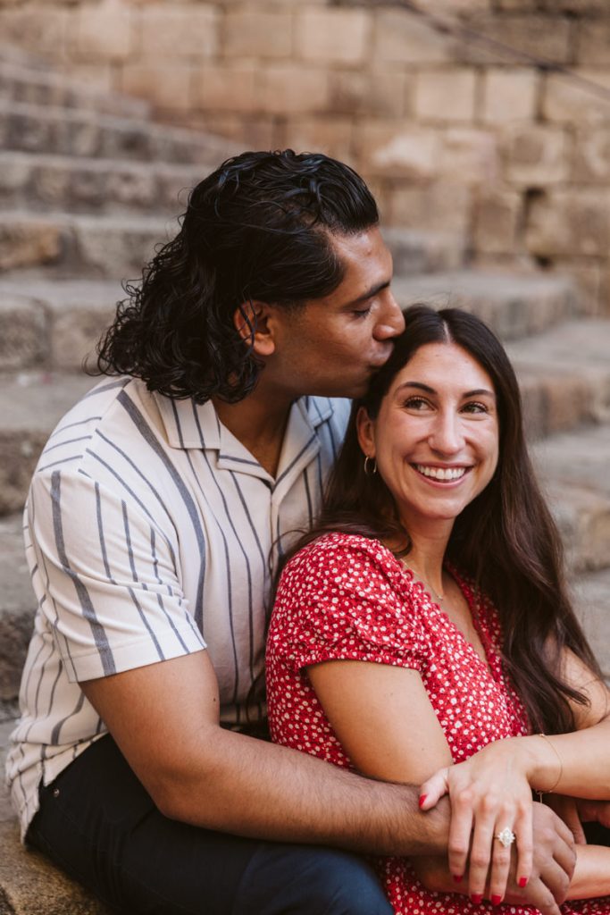 A man sits on stone steps and kisses a smiling woman on the head from behind. She wears a red patterned dress, and he wears a white shirt with dark stripes, both looking happy and relaxed.