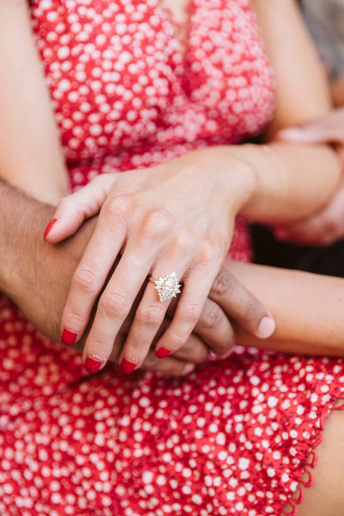 Close-up of a womans hand with red nail polish, wearing a diamond engagement ring. Her hand rests on another persons arm, and she is wearing a red, patterned dress.