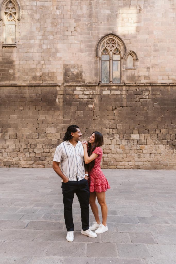 A smiling couple stands close together on a stone pavement in front of an old brick wall with arched windows. The woman in a red dress playfully holds the man’s shirt, while the man smiles with his hands in his pockets.