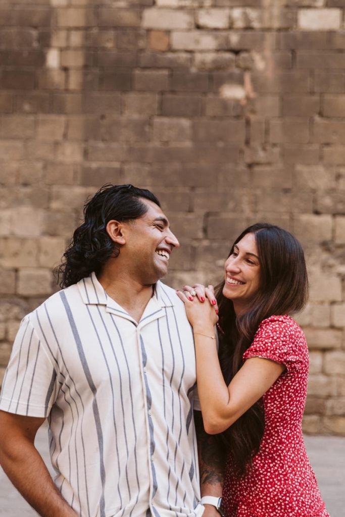 A smiling couple stands close together in front of a stone wall. The man wears a white shirt with dark stripes, and the woman in a red patterned dress leans on his shoulder, both looking at each other and laughing.