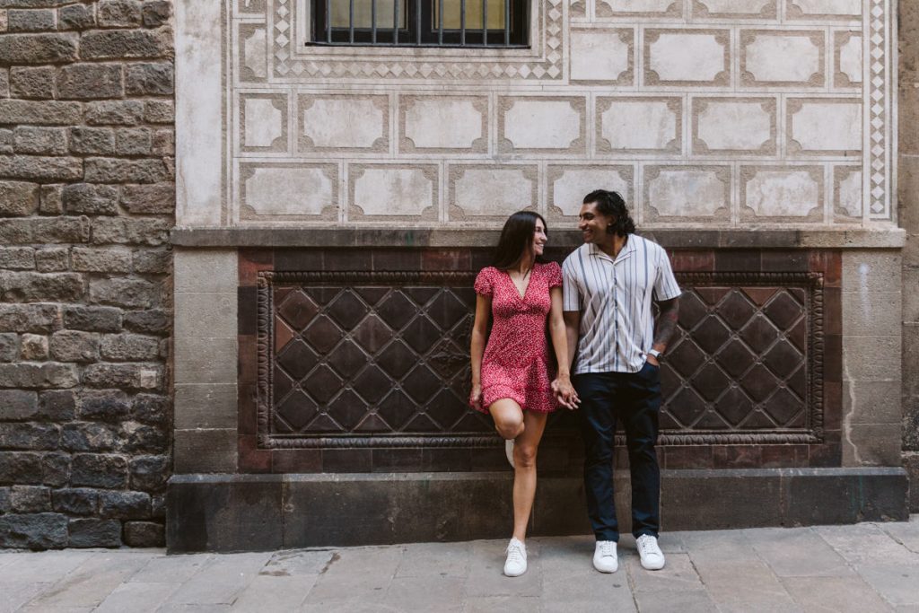 A couple stands holding hands and smiling at each other against a decorative stone wall, both dressed casually in summer clothes and white sneakers.