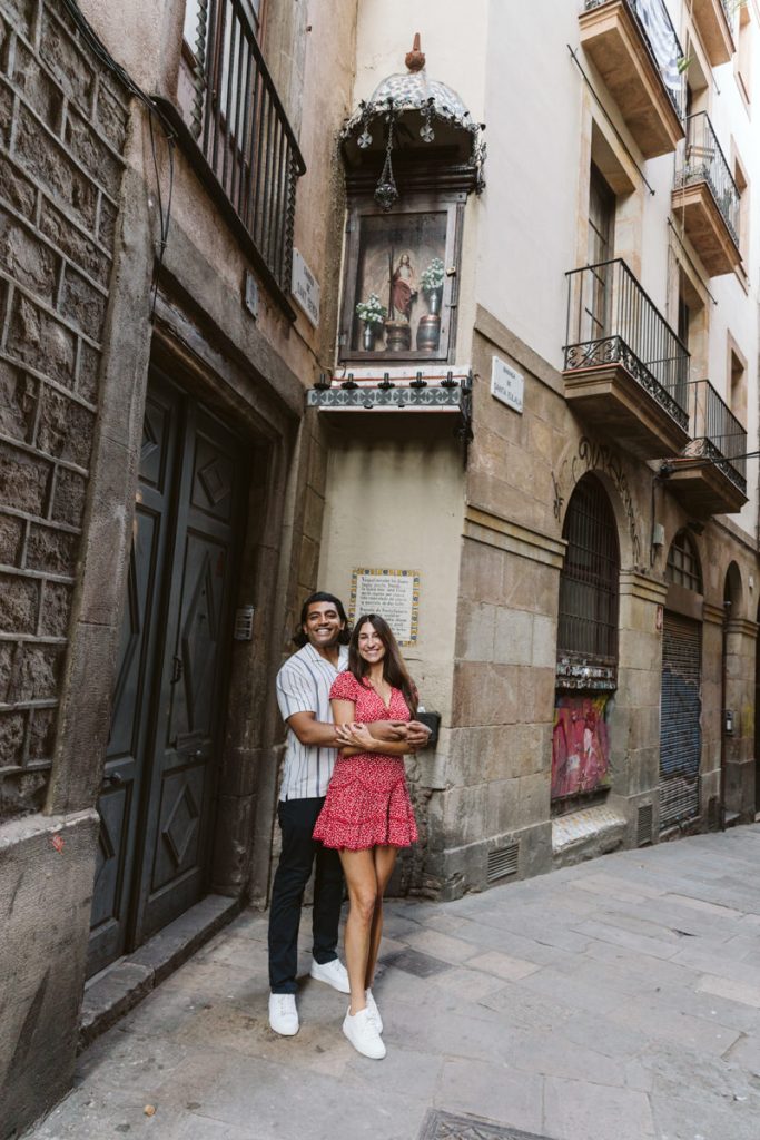 A couple stands smiling and embracing in a narrow European street, with tall buildings, balconies, and a religious shrine mounted on the wall above them. The woman wears a red dress and the man a striped shirt.